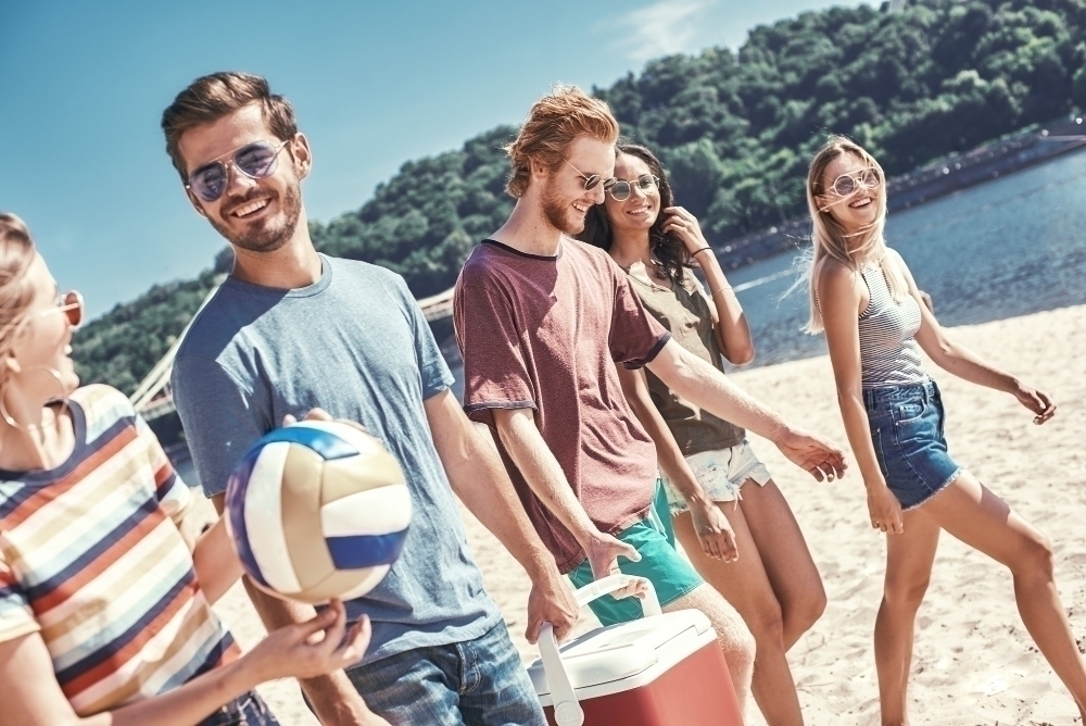 Cheerful young people walking by the beach to the sea while two men carrying plastic cooler