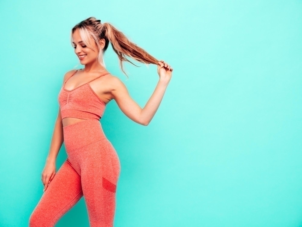 Sexy fitness smiling woman in pink sports clothing Young beautiful model with perfect bodyFemale posing near blue wall in studioCheerful and happy Holding her hair tail