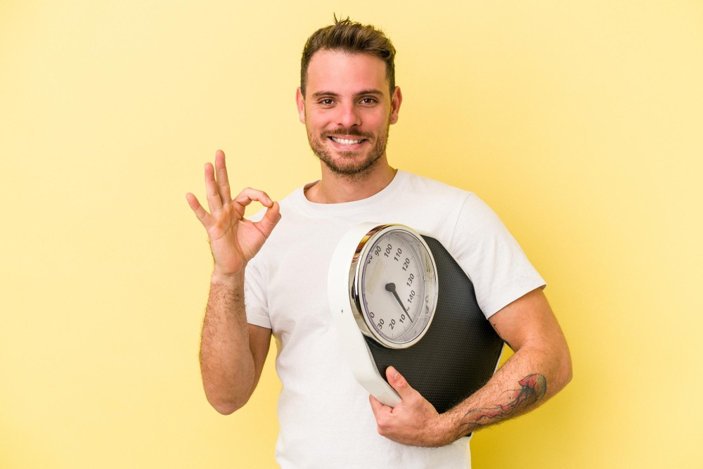 Young caucasian man holding a scale isolated on yellow background cheerful and confident showing ok gesture.