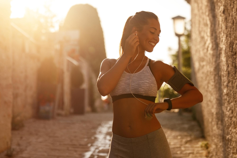 Young happy athletic woman using smart phone on her arm band and listening music while preparing to exercise in the city