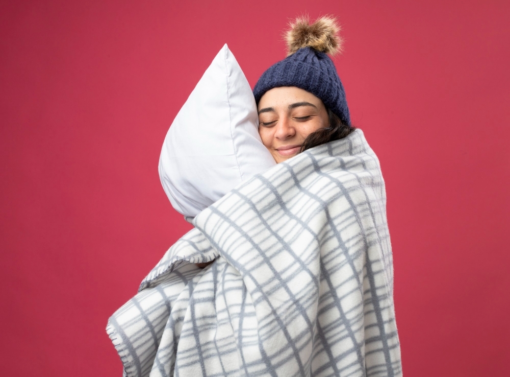 pleased young caucasian ill girl wearing robe winter hat scarf wrapped plaid standing profile view hugging pillow with closed eyes isolated crimson background with copy space